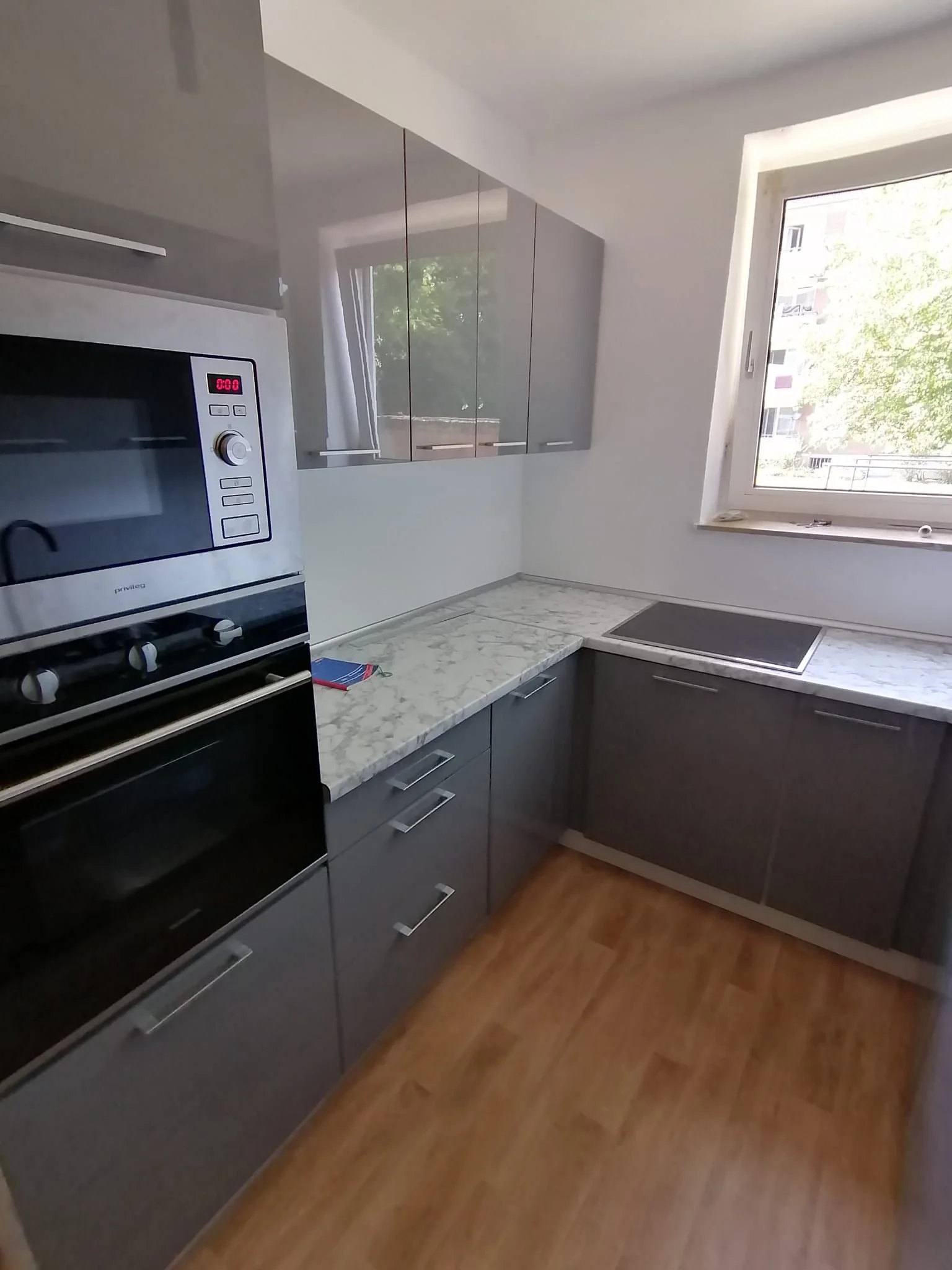Graue Küche mit heller Steinoptik-Arbeitsplatte, Fenster und Gerätehochschrank